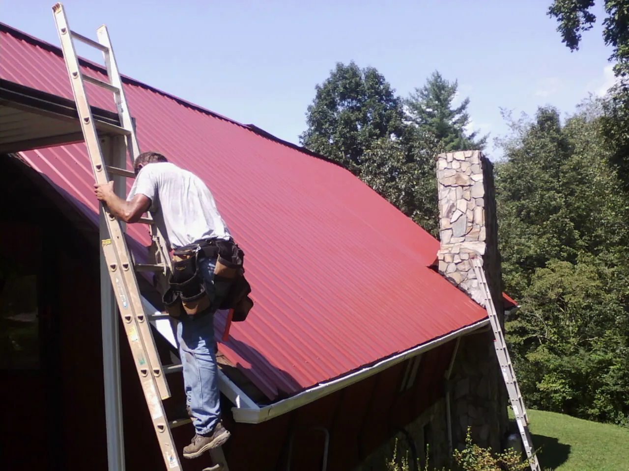 Roofer on a ladder beside a red metal roof and stone chimney — Soffit & Fascia Repair by Roofing Brothers in Winlock
